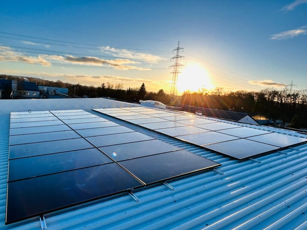 Solarmodule auf einem Wellblechdach bei Sonnenuntergang mit Strommast im Hintergrund.