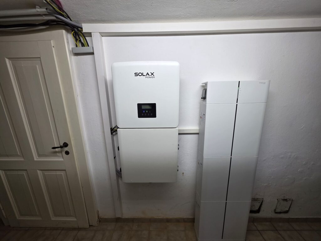 Weißes Solax-Gerät an der Wand montiert, daneben ein hohes, schmaleres, technisches Gehäuse auf hellem Fliesenboden.