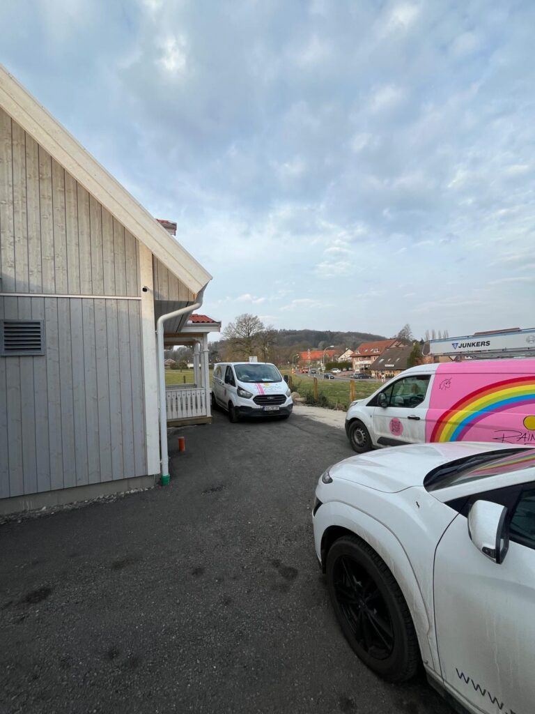 Drei geparkte Transporter neben einem hellgrauen Holzhaus unter bewölktem Himmel.