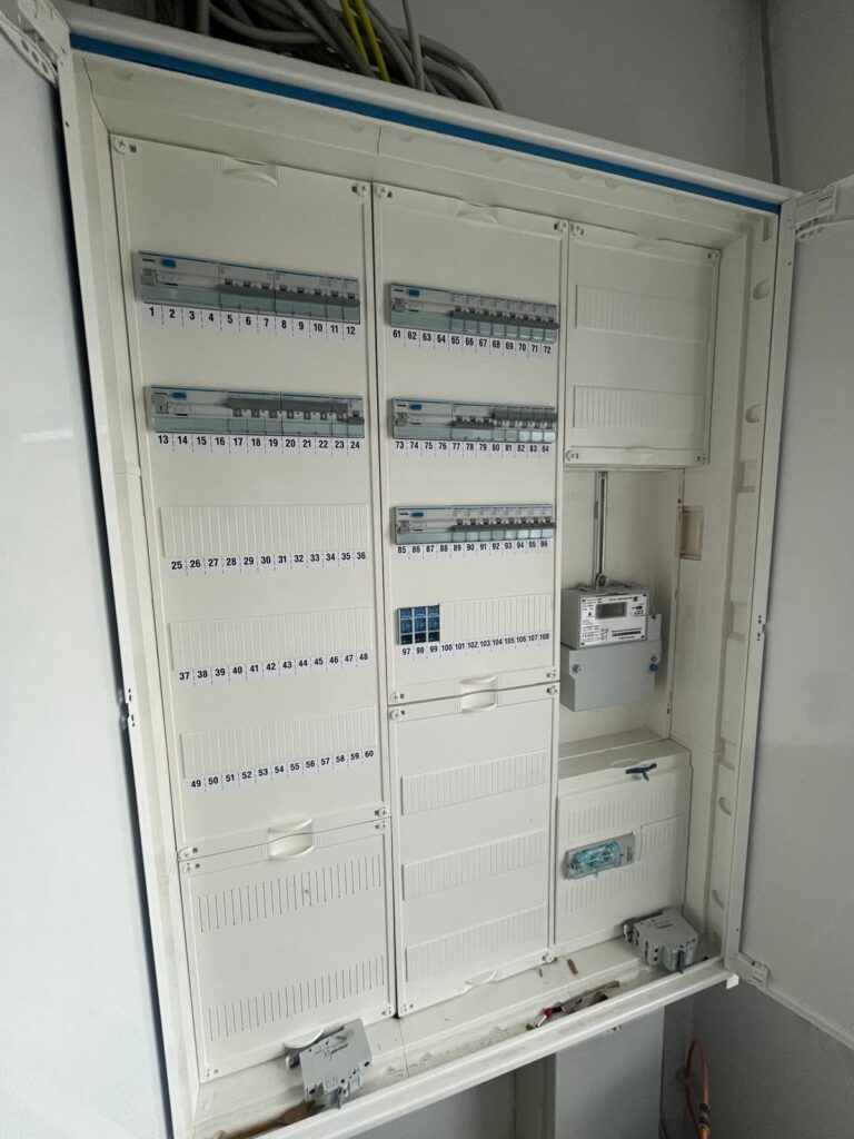 Geöffneter Verteilerkasten mit nummerierten Sicherungsautomaten und einem Zähler, darüber Kabelstränge.