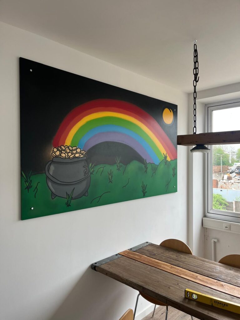 Wandgemälde mit Regenbogen und Topf voller Gold auf Wiese in einem Raum mit industriellem Tisch.