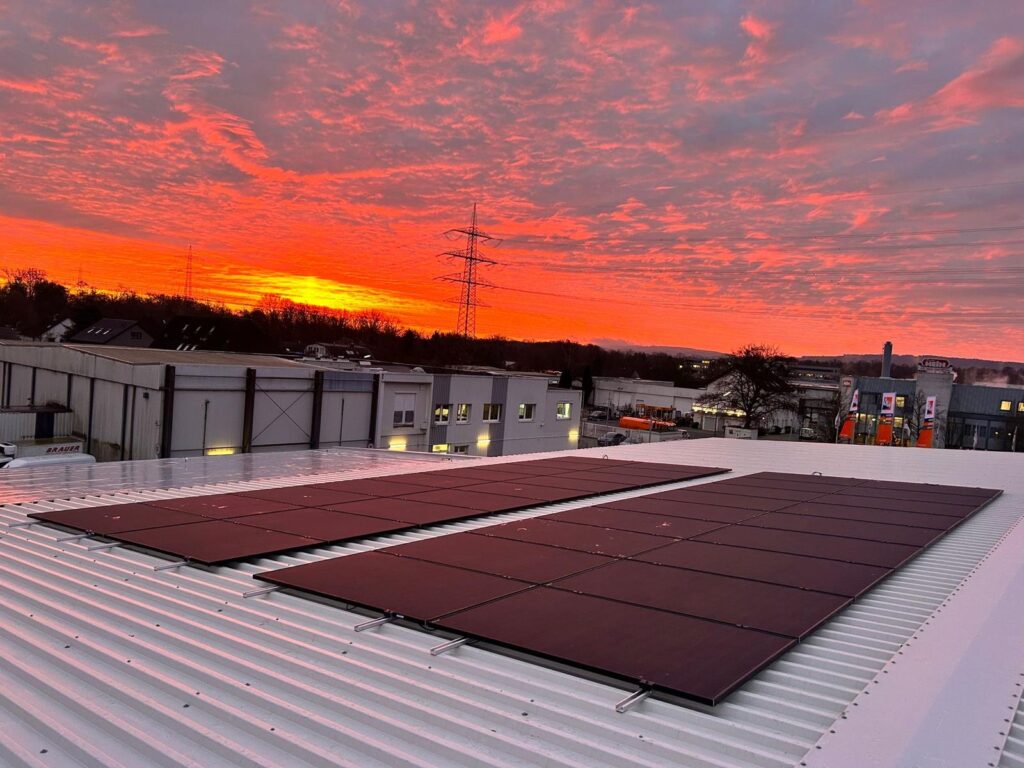 Photovoltaikmodule auf weißem Metalldach bei Sonnenuntergang, mit intensiv rot-orangefarbenem Himmel und Gebäuden im Hintergrund.