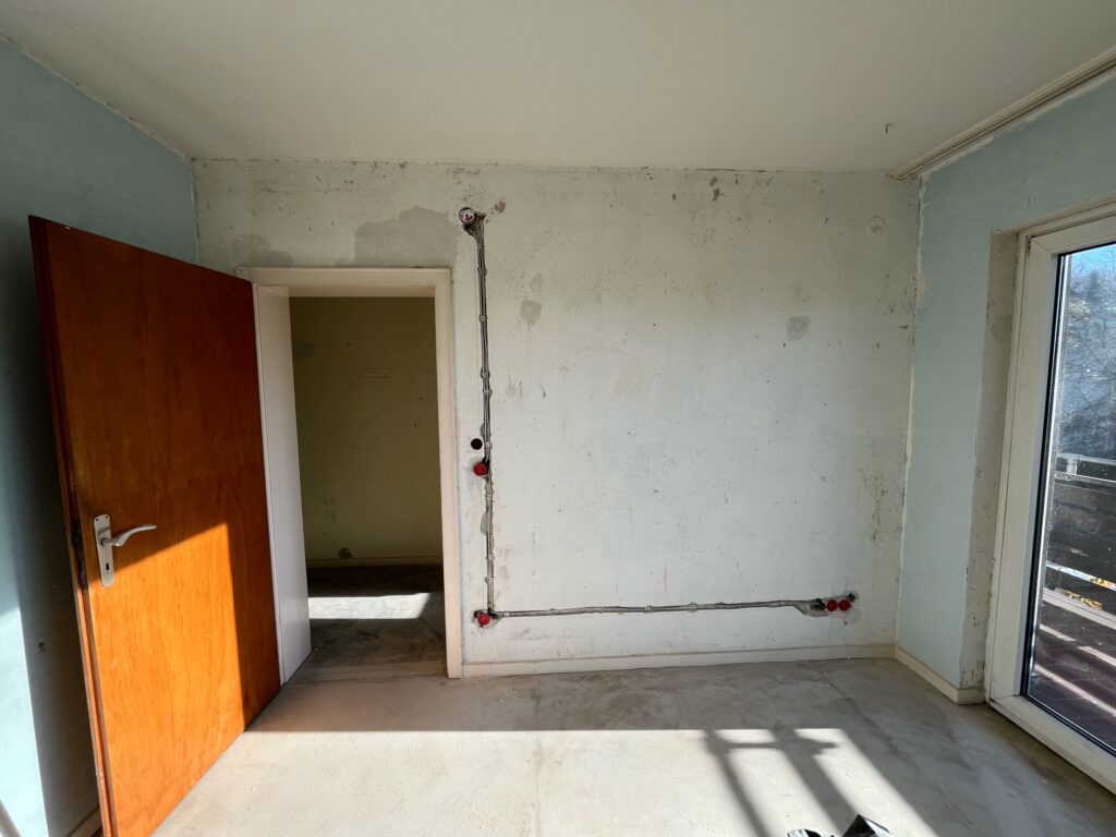 Leeres Zimmer mit offener Holztür, unverputzter Wand, sichtbarer Elektroinstallation und Fenster mit Balkonausblick.