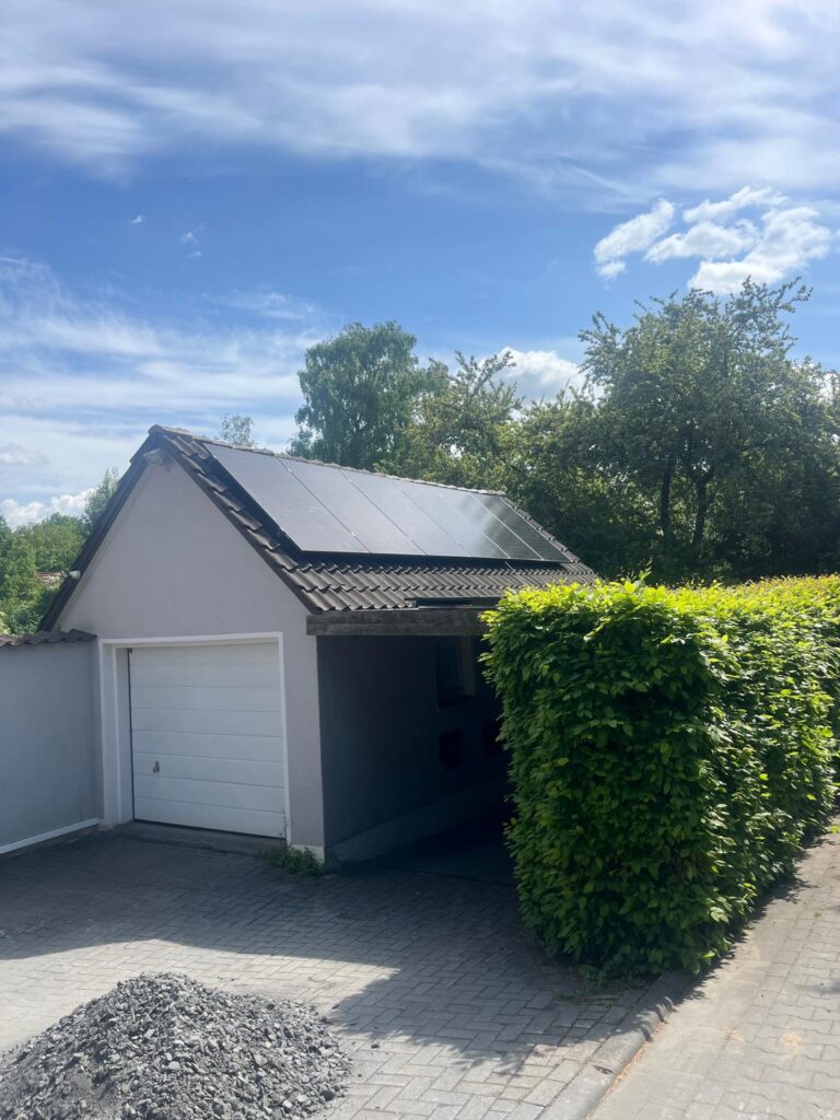 Graue Garage mit Solarpanelen auf dem Dach, grüne Hecke rechts, Schutthaufen auf gepflastertem Weg.