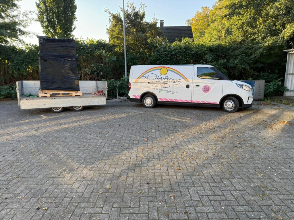 Weißer Lieferwagen mit Anhänger, beladen mit schwarzer, in Folie gewickelter Palette, steht auf gepflastertem Parkplatz.