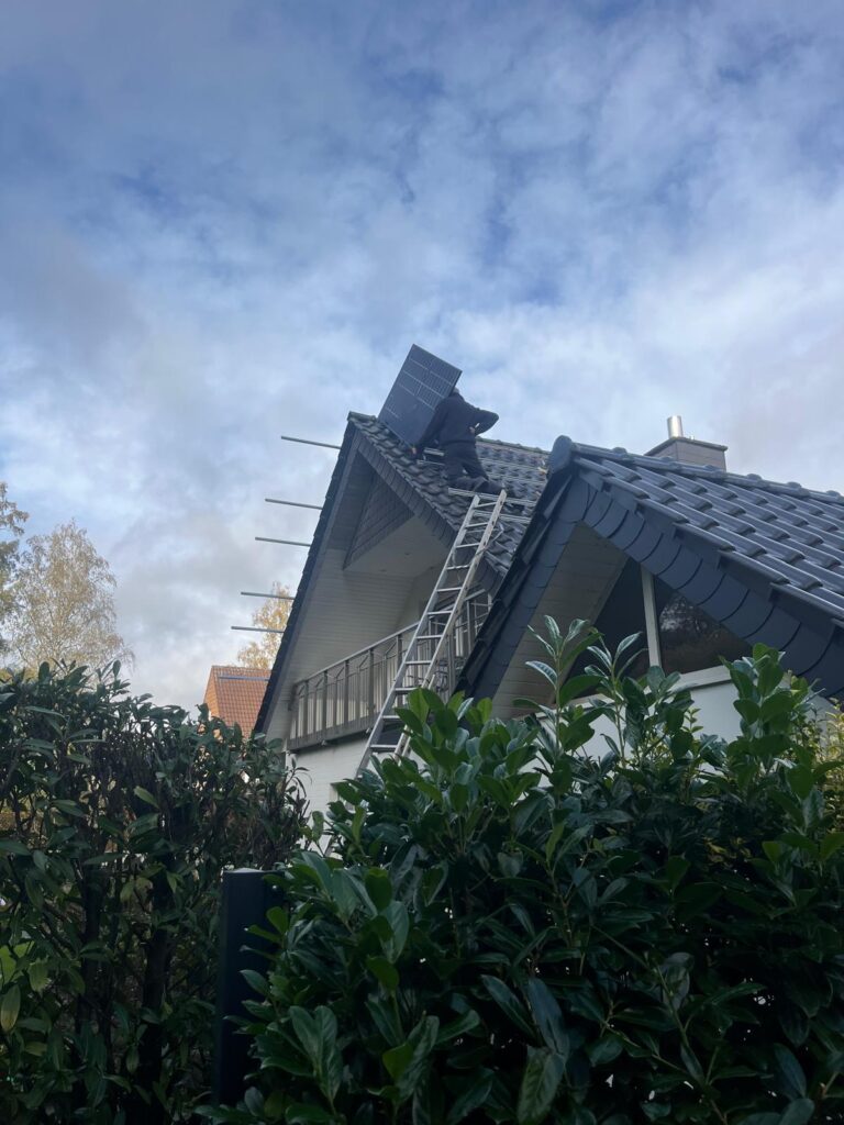 Person installiert Solarpanel auf Hausdach, Leiter angelehnt, bewölkter Himmel.