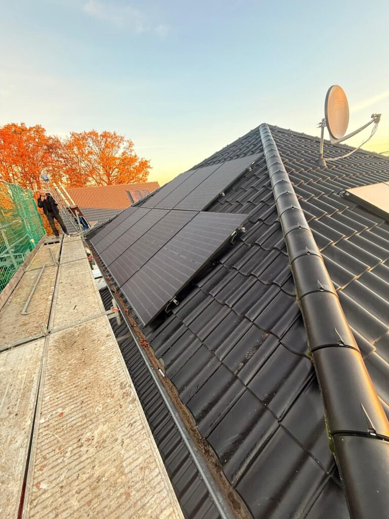 Schräge schwarz verkachelte Dachfläche mit Solarpaneelen und Satellitenschüssel, Gerüst links, herbstliche Bäume im Hintergrund.