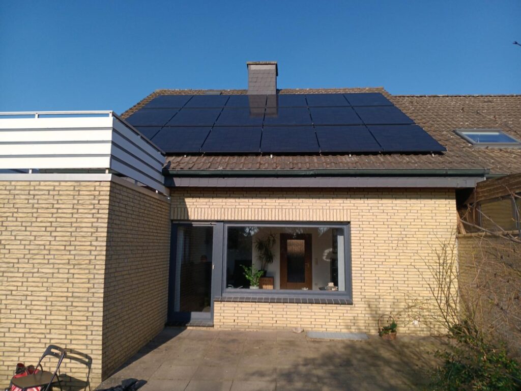 Flachdachhaus mit Solarpaneelen auf dem Ziegeldach, weißer Balkon, obere Etage, blauer Himmel.