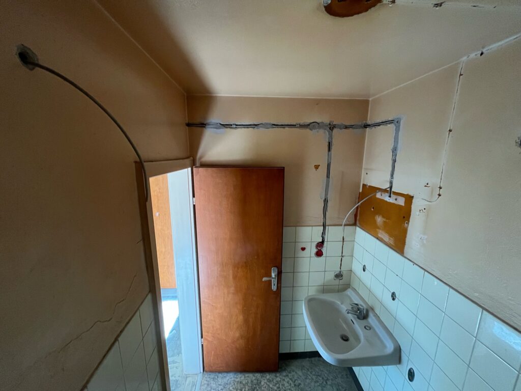 Kleines Badezimmer mit Waschbecken, Fliesenwand und sichtbarer Kabelinstallation oberhalb eines Holzfensters.