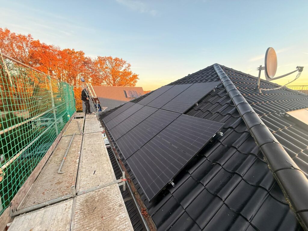 Dach mit schwarzen Solarpaneelen, daneben Gerüst mit Netzen, im Hintergrund Bäume mit Herbstlaub.