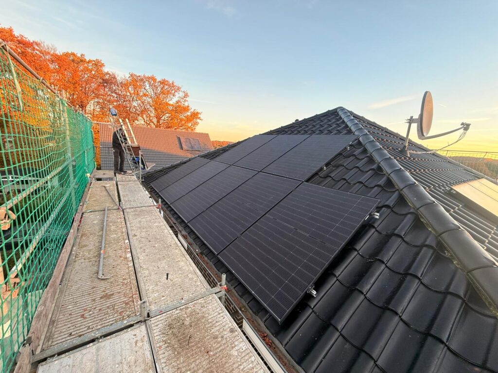 Dach mit Solarmodulen und Satellitenschüssel, Gerüst und Netz links, herbstliche Bäume im Hintergrund.