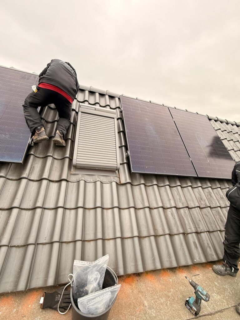 Person installiert Solarmodule auf Ziegeldach, neben Fensteröffnung; Akkuschrauber und Eimer im Vordergrund.