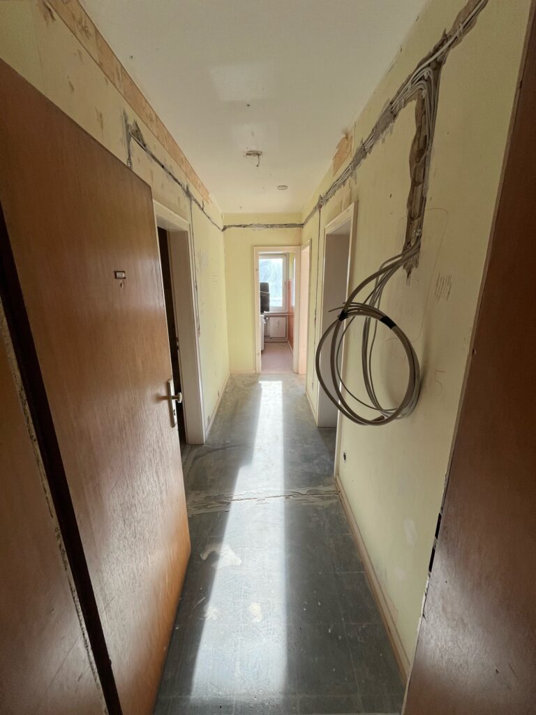 Renovierungsbedürftiger Flur mit offenen Elektrokabeln, teils abgerissener Tapete, Holztür, Sicht auf Fenster am Ende.