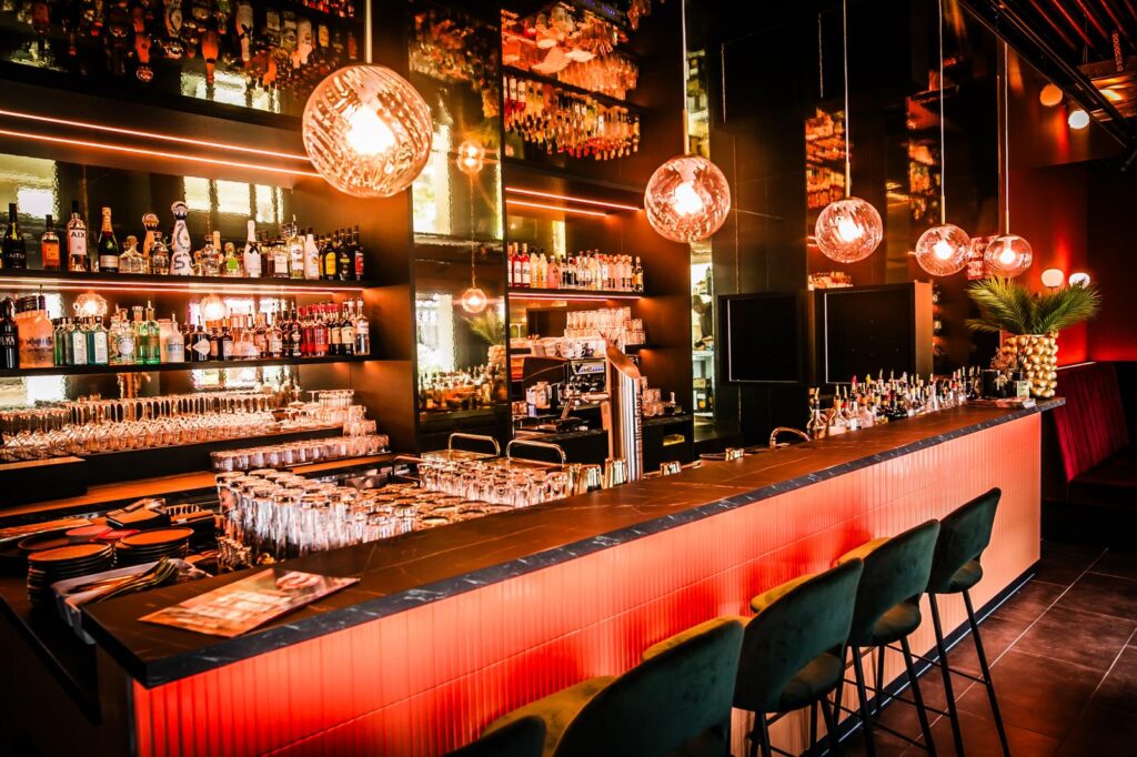 Modern eingerichtete Bar mit beleuchtetem Tresen, Gläsern, Spirituosenflaschen und hängenden, warmen Leuchten.