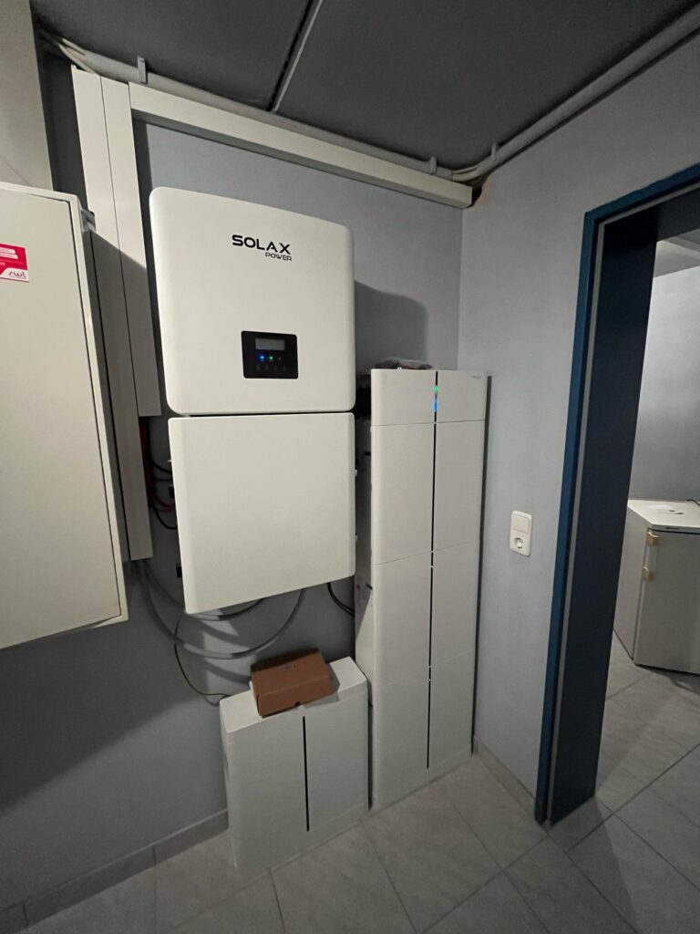 Weißes Solax Power-Energiemanagementsystem an der Wand montiert, darunter weiße Kästen und ein Karton.