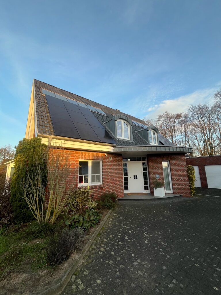 Zweistöckiges Backsteinhaus mit Solarpaneelen, weißer Eingangstür und Vorgarten, blauer Himmel im Hintergrund.